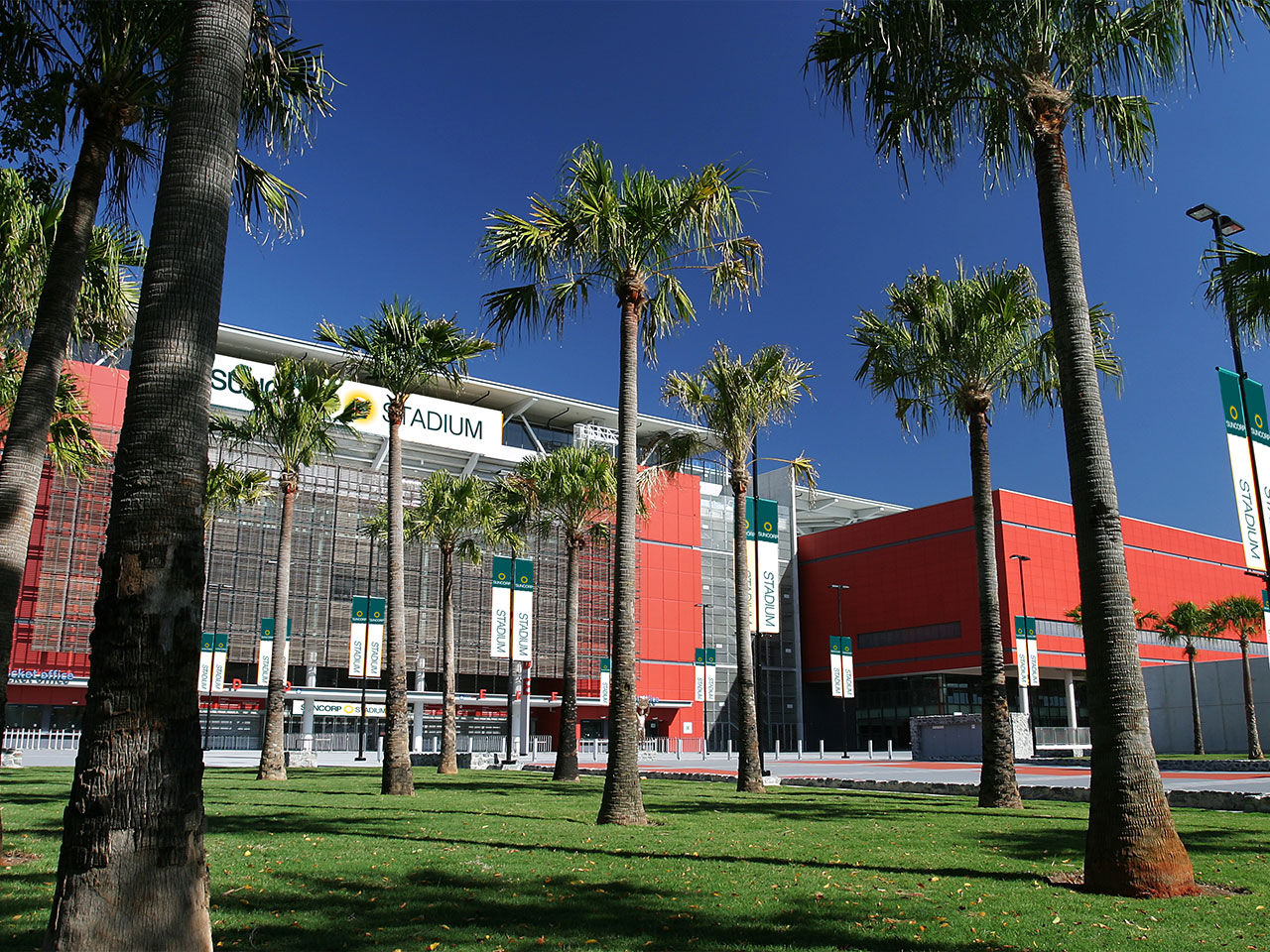 Suncorp Stadium's North Plaza. Palm trees and greenery in the foreground, stadium entrance in the background on a sunny day.