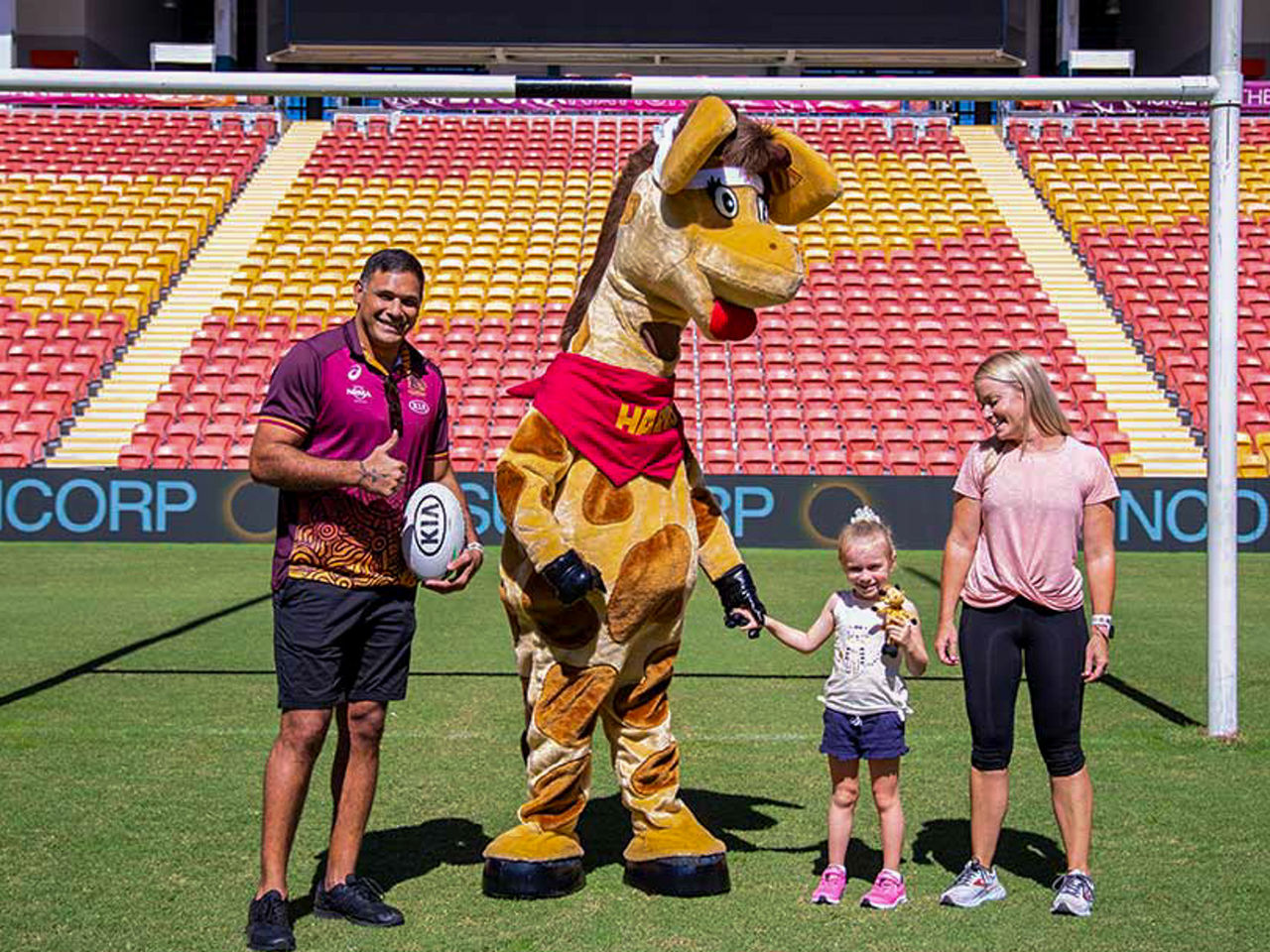 Image of Broncos star Justin Hodges (Far Left), Healthy Harold (Left), Lily Guy (Right) and Kate Maroulis (Far Right) stood on the field smiling at the camera. Justin Hodges is holding a ball whilst giving a thumbs up. Healthy Harold and Lily are holding hands. Lily is also holding a Healthy Harold stuffed toy.