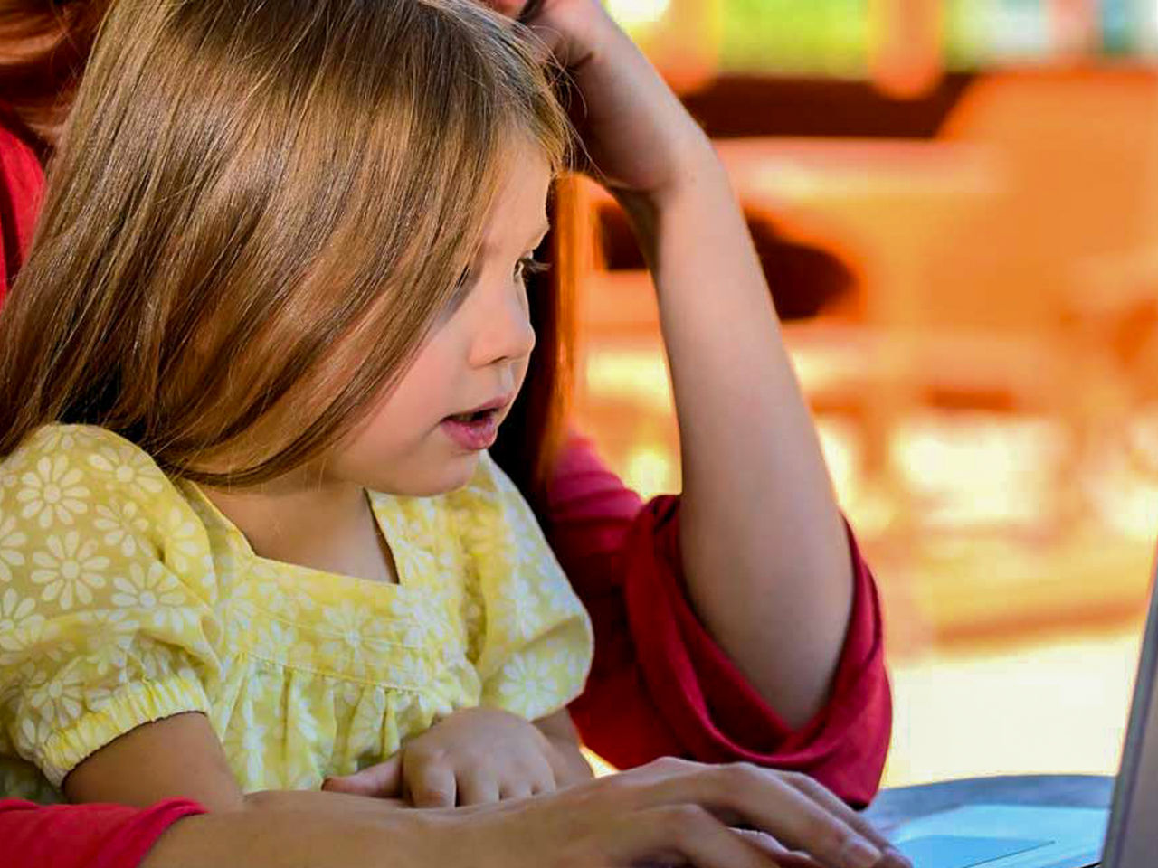 Image of a young person in a yellow flower outfit, sat on a person's lap, looking off camera at a laptop screen.