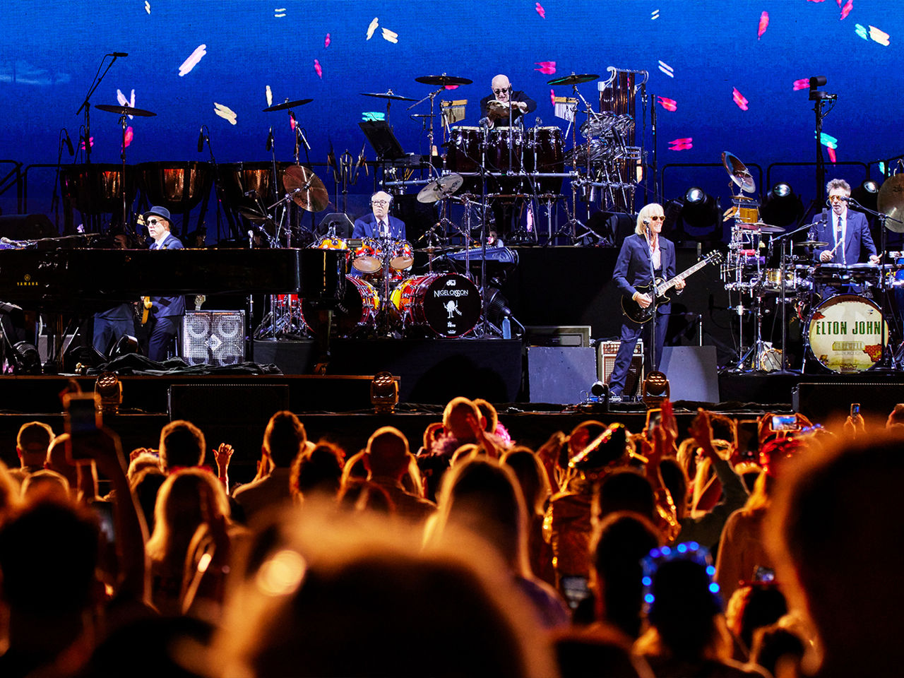 Crowd POV of the Elton John concert at Suncorp Stadium. The band is on the stage and confetti graphics are on the big screen behind them.