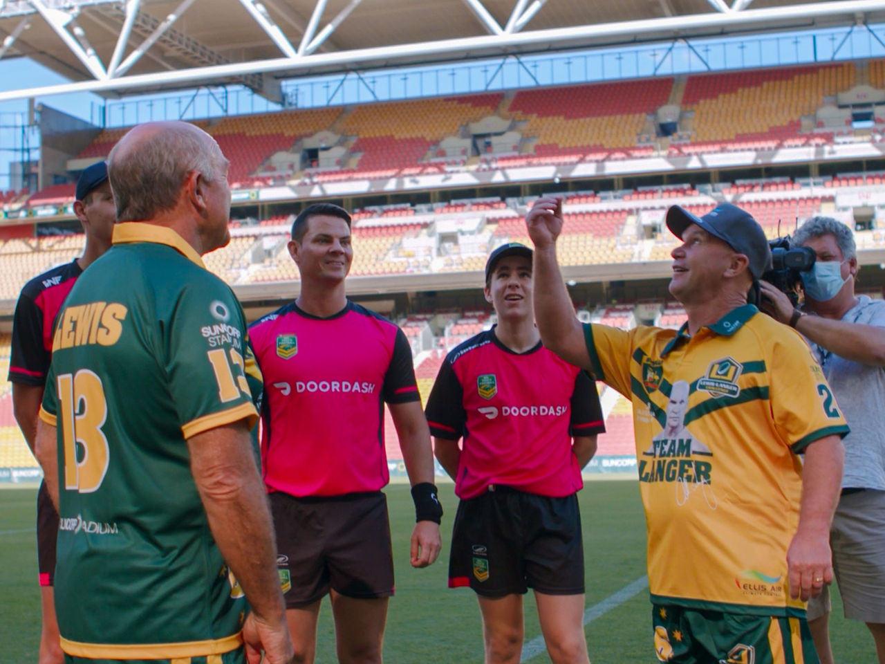 Image of Wally Lewis (Left), referees (Middle) and Alfie Langer (Right) on Suncorp Stadium field. Alfie Langer is mid coin toss. There is a camera man filming to the far right.
