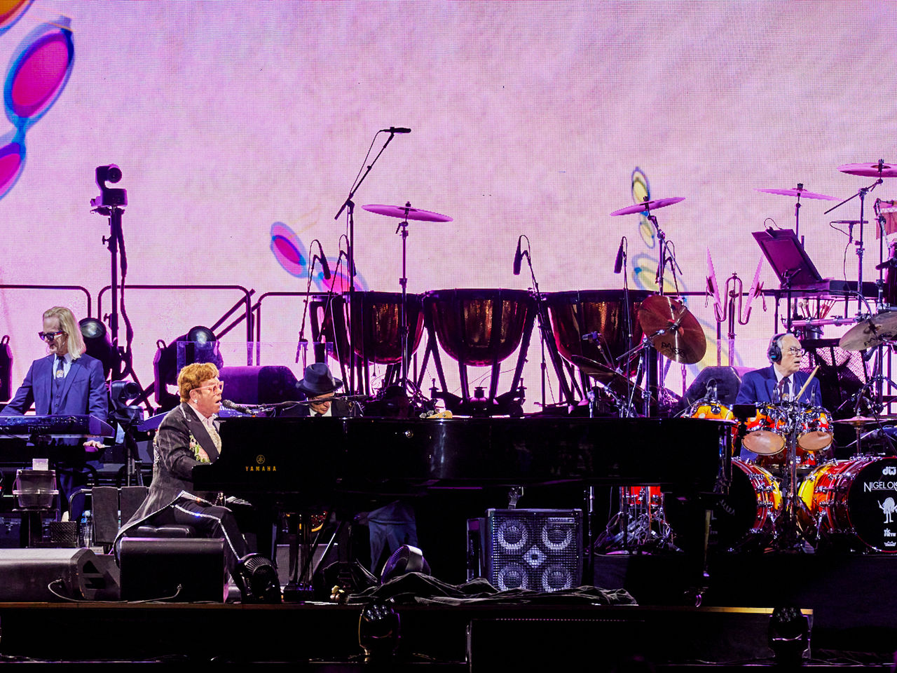 Image of Elton John singing into a microphone whilst sat at his piano on stage.