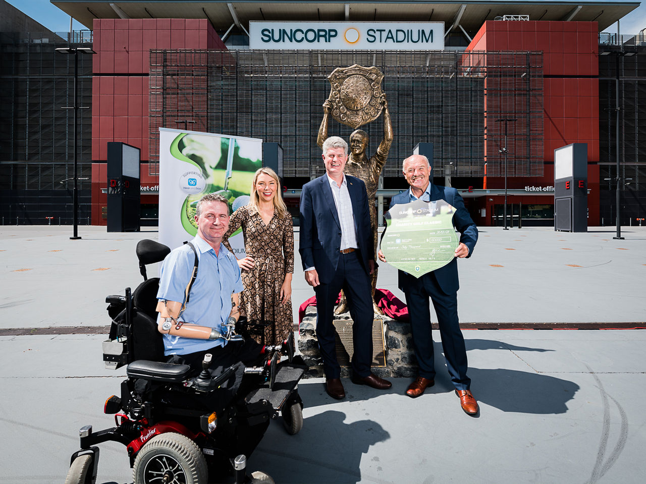 Image of (Left to Right) Matthew Ames, Jonty Bush, Sports Minister Stirling Hinchliffe and Wally Lewis, in front of the Wally Lewis Statue with Suncorp Stadium behind them. Wally is holding a sign.