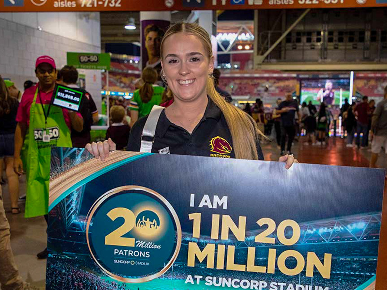 Image of Petrea Spencer, the 20th Millionth Patron, in Suncorp Stadium, holding a sign that says 'I am 1 in 20 Million at Suncorp Stadium' and smiling at the camera.