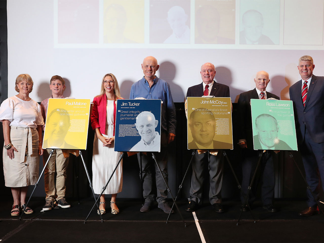 Image of Suncorp Stadium's Newest Media Hall of Fame Inductees stood on a stage. Pictured left to right, Paul Malone's wife and son, Local State Member for Cooper Jonty Bush, Jim Tucker, John McCoy and Ross Lawson. They are all smiling at the camera whilst stood behind their respective plaques.