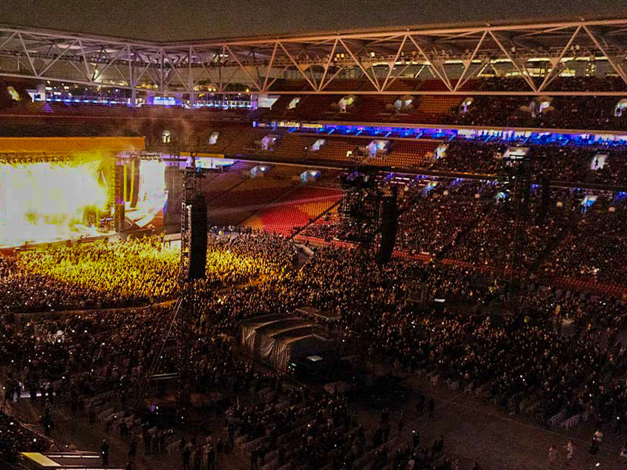Image of a concert at Suncorp Stadium. It shows the bright stage in the far left side and the crowd of people in attendance.
