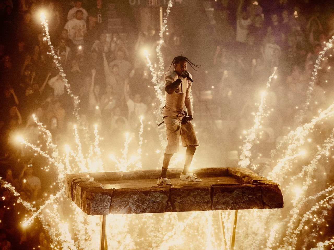 Travis Scott mid-performance on a elevated stage with pyrotechnics going off around him.