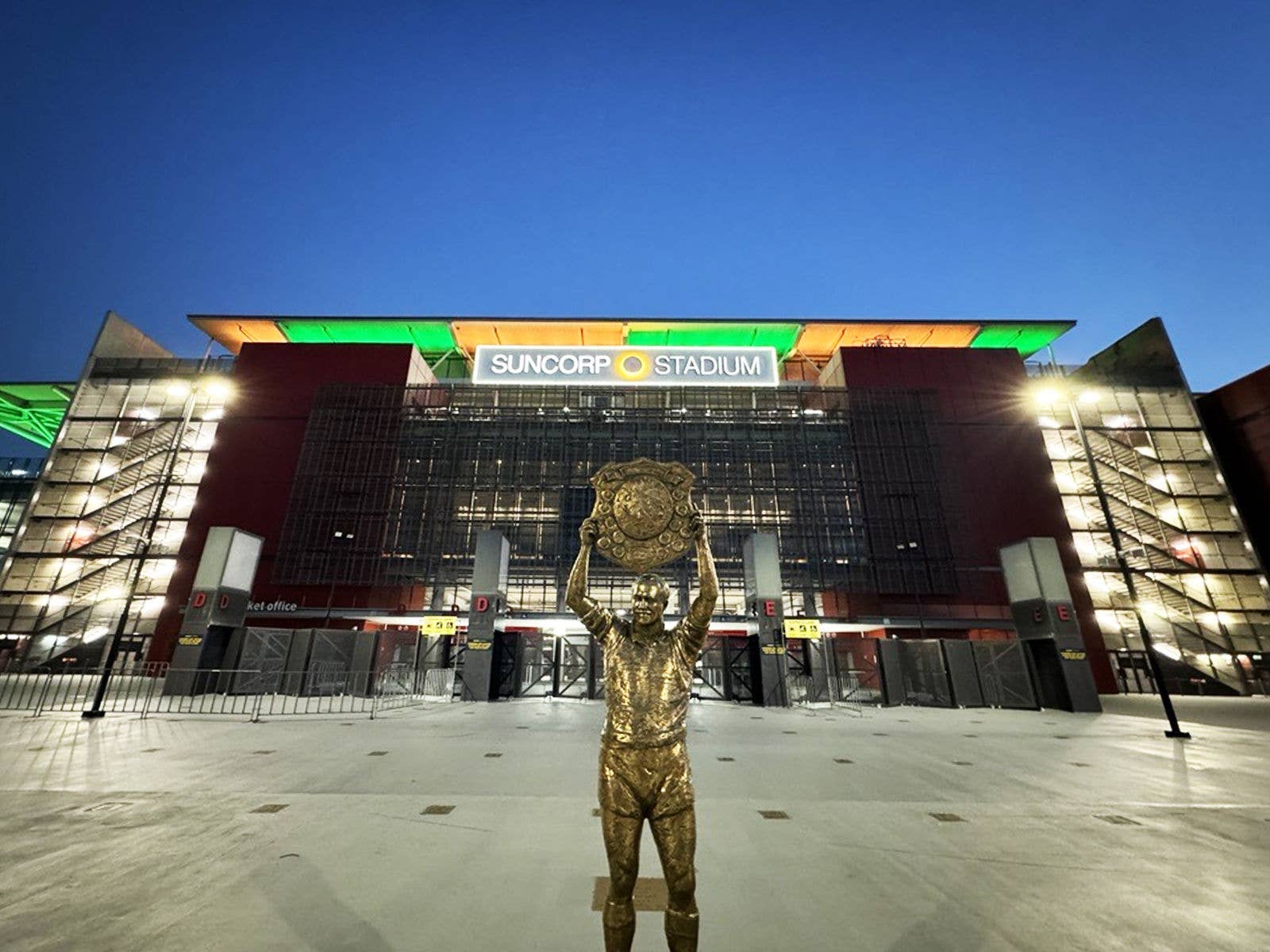 The Wally Lewis Statue with Suncorp Stadium lit up green and gold in the background