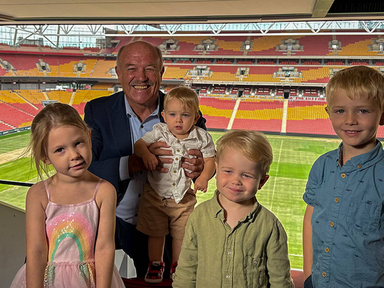 Suncorp Stadium's Biggest Donation Yet a Hole-In-One for Hear and Say. Image of Wally Lewis and Hear and Say Family Members with Suncorp Stadium field in the background. From Left to Right: Isla (3 years), Wally Lewis, Archie (16 months), Finn (3 years), Elias (5 years).
