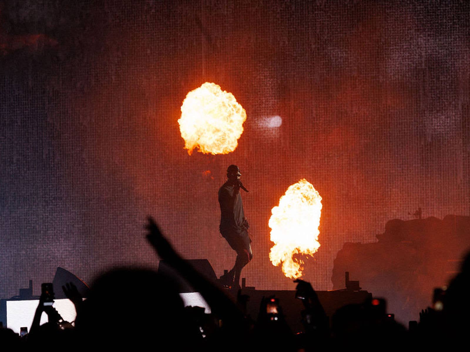 Travis Scott performing on stage at Suncorp Stadium in 2024 with pyrotechnics going off behind him. Fans arms can be seen in the foreground.