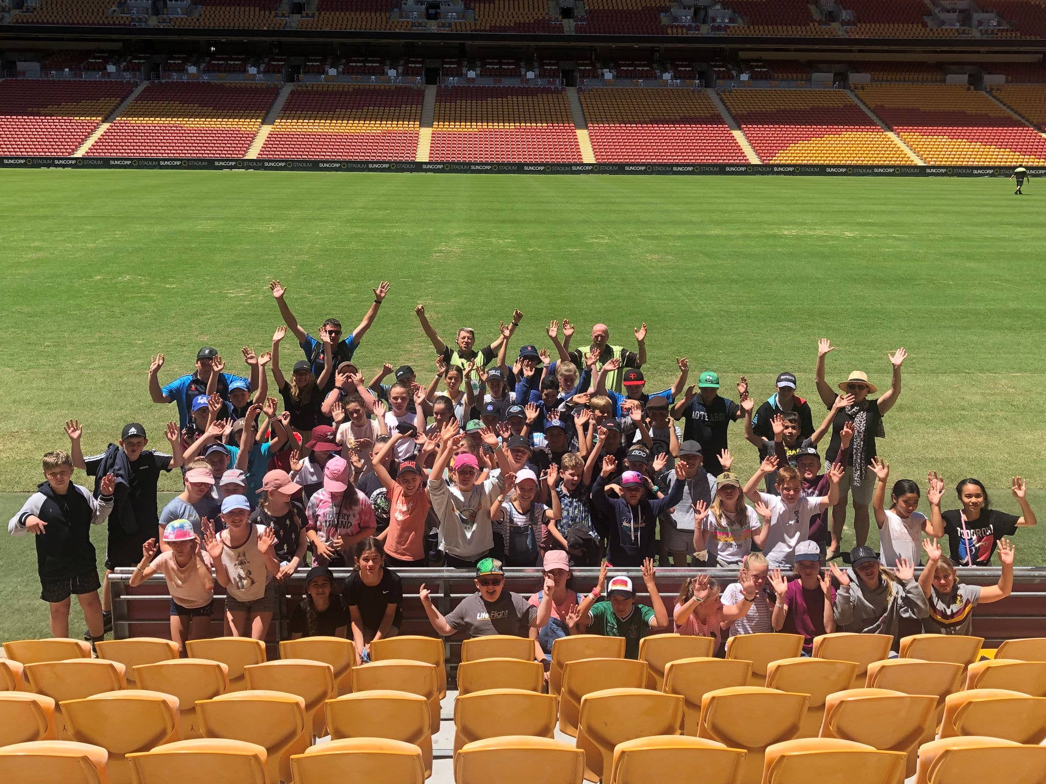 Group of people smiling up at the camera with their hands in the air whilst on the Suncorp Stadium sideline during their tour visit.