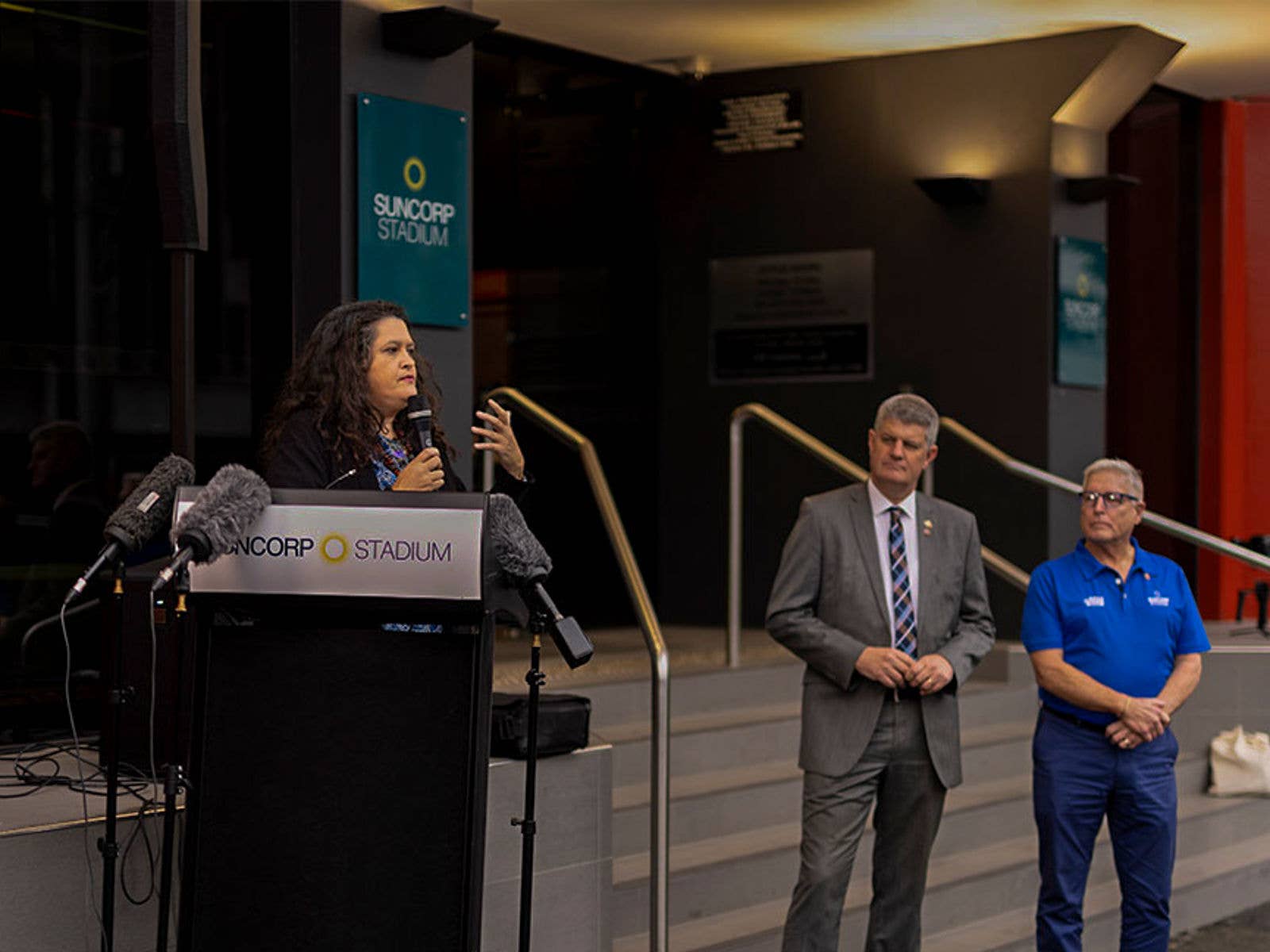 Artist speaking at 'Welcome' artwork presentation. Artist speaking into microphone at 'Welcome' artwork presentation in front of Suncorp Stadium. General Manager Alan Graham and Sports Minister are watching in the background.