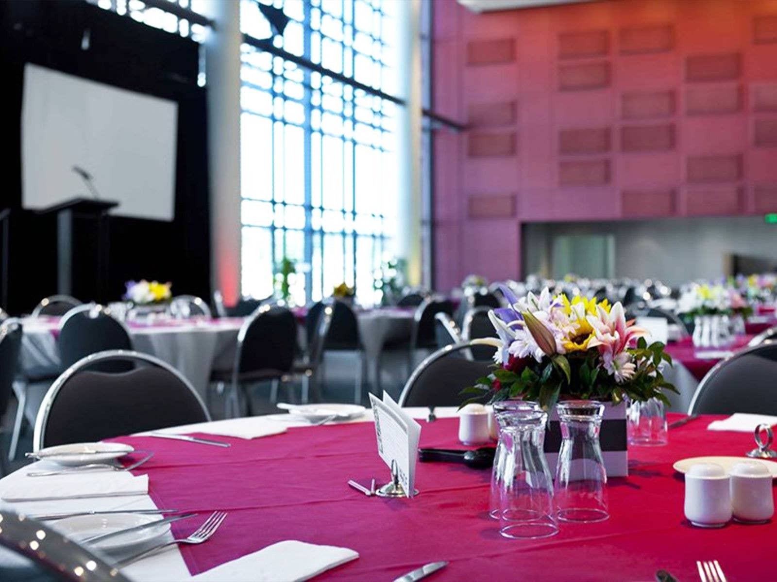 Internal view of function space at Suncorp Stadium. Circular tables are dressed with dishware and floral centre pieces. There is a stage with a large screen and microphone podium to the far left.