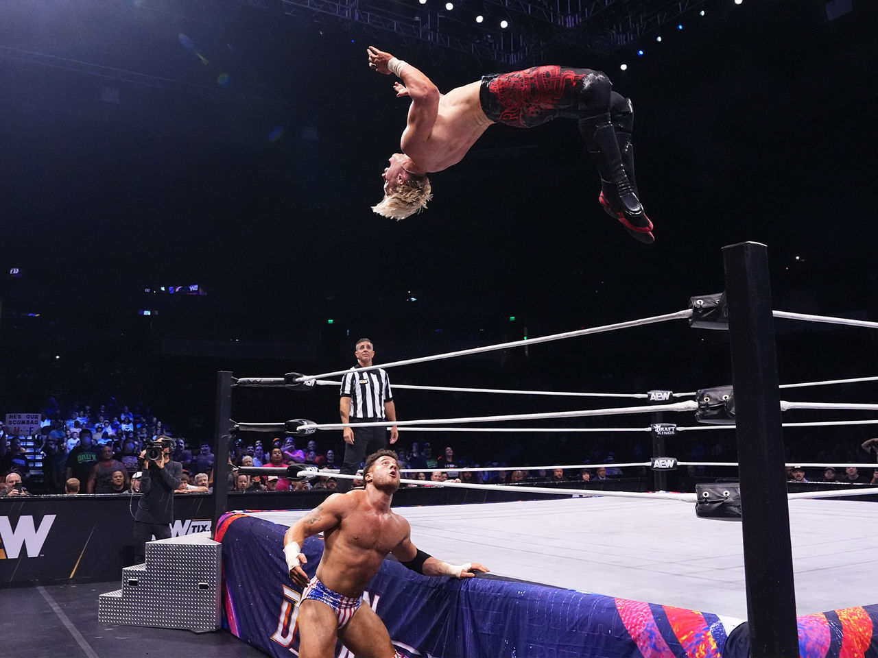 Image of two wrestlers mid fight. One is in the air, mid back flip from jumping off the ropes. The other is stood outside the ring on the ground looking up.