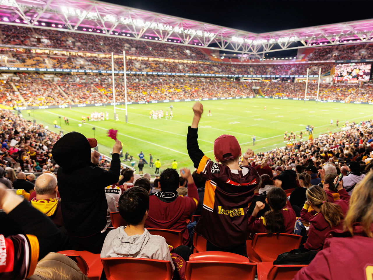 Image of Suncorp Stadium at nighttime during a Broncos game. The stadium is packed with fans. In the foreground of the image are two fans stood up in their seats with their hands in the air.